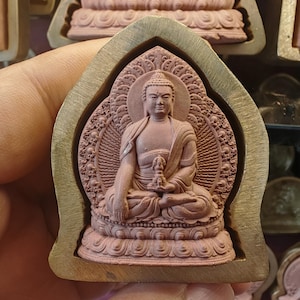 May include: A reddish-brown Buddha statue in a seated pose, set within a gold-coloured metal frame. The statue has detailed carvings and is set against a blurred background.