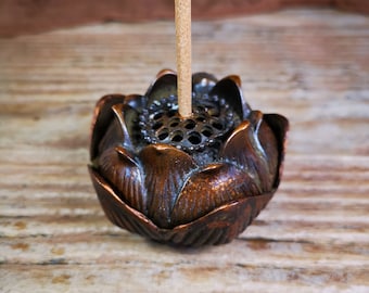May include: A bronze lotus flower incense holder with a brown incense stick inserted into the top. The holder has a decorative design and a small hole in the center.