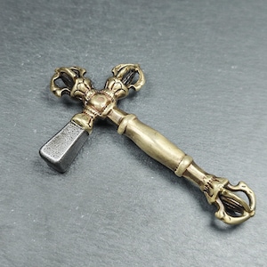 May include: An ornate metal vajra, a ritual object with a cross-shaped design. The handle and decorative elements are gold-toned, while the head is a darker, silver-grey colour. The vajra is presented on a dark grey surface.
