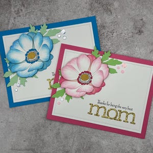 May include: Two handmade greeting cards with floral designs. One card has a blue border and a blue and white flower. The other card has a pink border and a pink flower. The text on the pink card reads "Thanks for being the very best MOM."