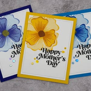 Pode incluir: Três cartões de Dia das Mães feitos à mão. Cada cartão apresenta um grande design floral em azul ou amarelo, com o texto "Happy Mother's Day" em uma fonte cursiva. Os cartões têm bordas coloridas e lantejoulas decorativas.
