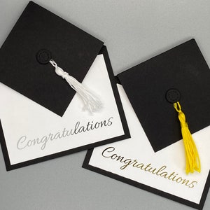 May include: Two black graduation cap shaped cards with white and gold tassels. The cards have white backgrounds with black borders and the word "Congratulations" written in silver and gold lettering.