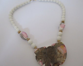 May include: White beaded necklace with a large, half-moon pendant. The pendant features a floral design with brown, pink, and gold accents. Additional pink and gray accents are on the necklace. The necklace is secured with a gold clasp.