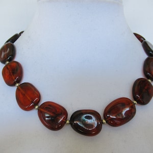 May include: A necklace featuring large, oval-shaped beads in shades of amber and dark brown. The beads are strung together with small silver-colored connectors, creating a bold and eye-catching accessory. The necklace is displayed on a white mannequin.