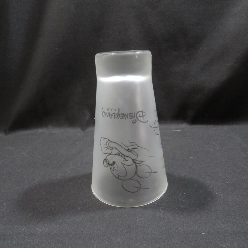 Op de afbeelding: Matglazen Disneyland Parijs souvenirbeker. De beker heeft een zwarte omtrek van Mickey Mouse en het Disneyland Parijs-logo. De beker heeft een uitlopende vorm en is enigszins doorschijnend.