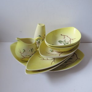 Può includere: Un servizio di piatti vintage in ceramica gialla con una crema, un saliera e diversi piatti e ciotole. Il set presenta un design a ramo marrone e foglie verdi. I piatti e le ciotole hanno una forma allungata.