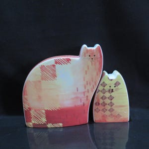 May include: Two ceramic cat figurines, one larger than the other. The larger cat is red, yellow, and white with a plaid pattern. The smaller cat is yellow, red, and white with a diamond pattern. Both cats have black eyes and whiskers.