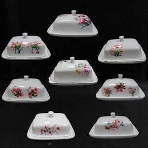 May include: A selection of white ceramic butter dishes with lids, each decorated with a different floral pattern. The lids display various flower arrangements in pink, red, yellow, and blue, set against a white backdrop.