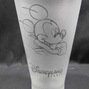 Op de afbeelding: Een mat glas met een schets van Mickey Mouse in beweging. Het ontwerp bevat het Disneyland Paris-logo. Het glas heeft een taps toelopende vorm en een gladde, matte afwerking, ideaal voor drankjes. Ongeveer 15 cm hoog.