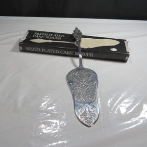 Può includere: Pala per torte argentata con un ornato motivo floreale sulla lama. La pala ha un manico decorativo ed è esposta con la sua scatola originale in bianco e nero. La scatola recita "Silver-Plated Cake Server".