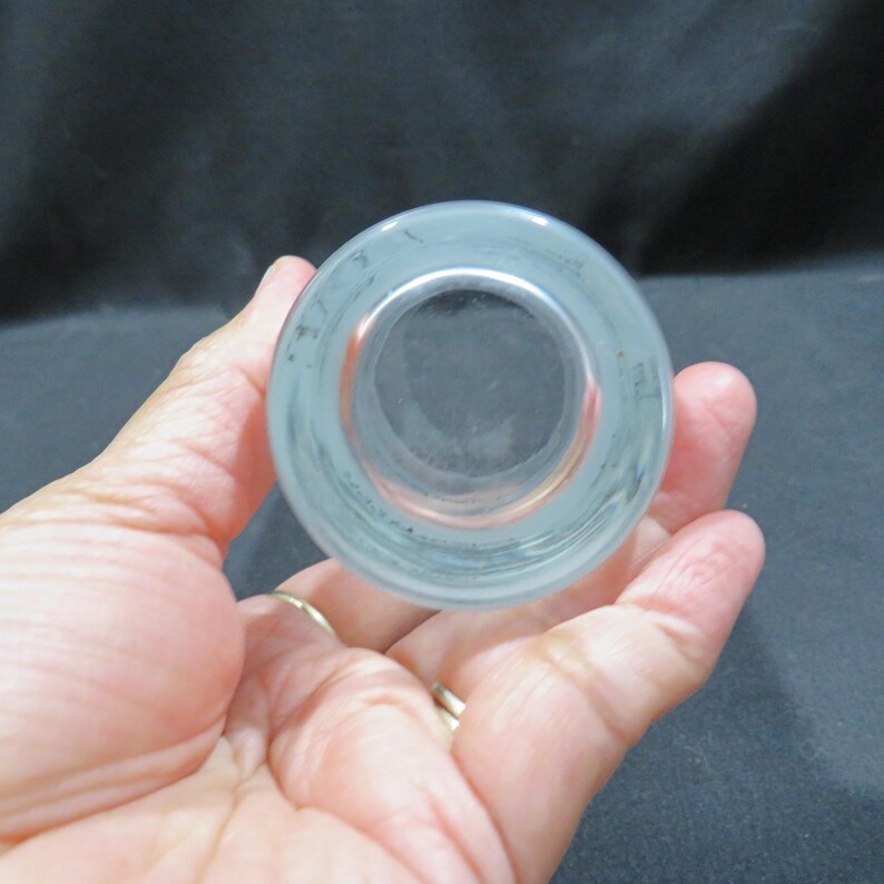 Op de afbeelding: Een helder glazen object met een cirkelvormige opening, vastgehouden in een hand. Het glas heeft een matte uitstraling en een donker interieur. Het object is waarschijnlijk een drinkglas of een kleine container.