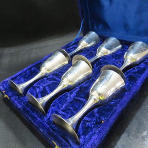 Pode incluir: Conjunto de seis cálices de prata com desenhos florais gravados, apresentados numa caixa forrada a veludo azul. Cada cálice tem uma haste fina e uma taça larga e rasa. Os cálices têm cerca de 13 cm de altura.