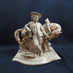 Könnte beinhalten: Keramikfigur, die eine Person in historischer Kleidung mit einem Pferd darstellt. Die Figur ist hauptsächlich cremefarben mit goldenen Akzenten auf dem Pferd, dem Hut und der dekorativen Schale. Der Sockel ist rechteckig.