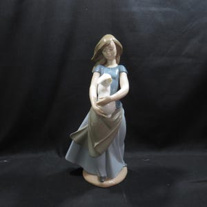 Peut inclure: Une figurine en porcelaine représentant une personne aux cheveux bruns vêtue d'un haut bleu et d'une longue jupe bleu clair avec un tablier beige. La figurine tient un petit lapin blanc dans ses bras et repose sur une base beige lisse et brillante.