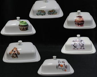 May include: Set of five white ceramic butter dishes with lids. Each dish features a different design: a truck with flowers, a cupcake, a dog, a cat, and two cows. The dishes are perfect for serving butter, cheese, or other small snacks.
