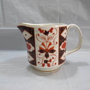 May include: Cream-colored ceramic pitcher with a decorative floral and geometric pattern in shades of red and brown. The pitcher has a curved handle and a spout, with gold trim along the rim and base. The design features stylized flowers and diamond shapes.