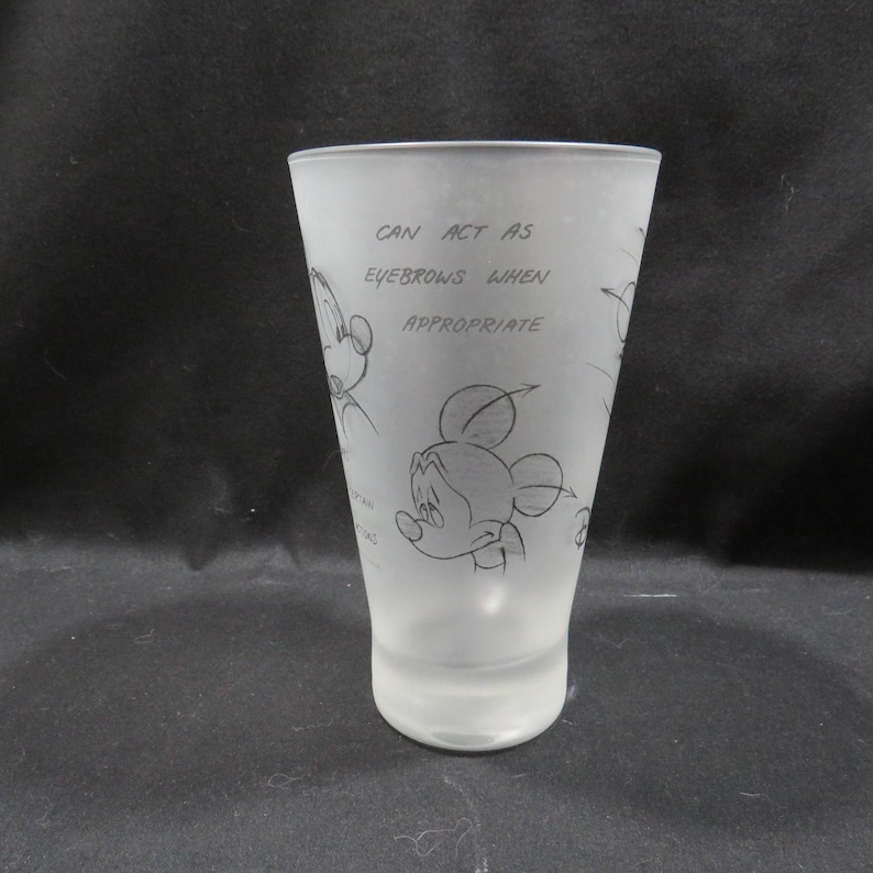 Op de afbeelding: Matte glazen tumbler met schetsen van Mickey Mouse en de tekst "CAN ACT AS EYEBROWS WHEN APPROPRIATE." De cilindervormige beker is geschikt voor drankjes. Het ontwerp is een speelse verwijzing naar het beroemde stripfiguur.