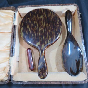 Op de afbeelding: Een vintage toilettafelset met een handspiegel, haarborstel en een kleine borstel, allemaal met een schildpadpatroon. De set wordt gepresenteerd in een bruine, imitatie leren koffer met een crèmekleurige satijnen voering. De spiegel is ongeveer 13 cm in diameter.