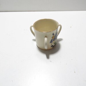 Op de afbeelding: Een klein, wit keramiek kopje met twee handvatten. Het kopje heeft een gouden rand en een kleurrijk ontwerp aan de zijkant.
