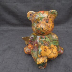 May include: A ceramic teddy bear figurine with a floral pattern. The bear is sitting with its legs crossed and has a gold tag on its foot.