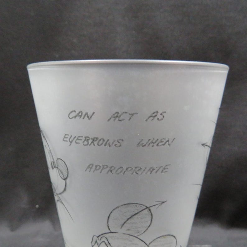 Op de afbeelding: Een matglas met een cartoontekening van Mickey Mouse. De tekst op het glas luidt "CAN ACT AS EYEBROWS WHEN APPROPRIATE". Het glas heeft een licht taps toelopende vorm.