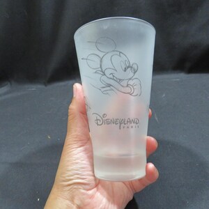 Op de afbeelding: Matte glazen tumbler met een schets van Mickey Mouse in beweging. De woorden "Disneyland Paris" staan eronder gedrukt. Het glas wordt in een hand gehouden.