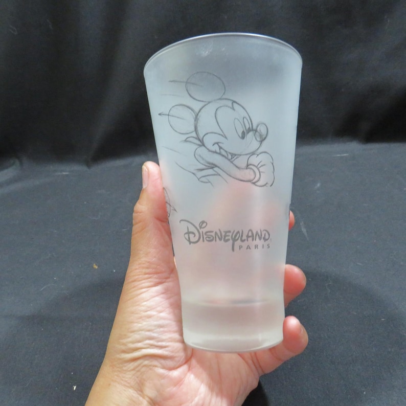 Op de afbeelding: Matte glazen tumbler met een schets van Mickey Mouse in beweging. De woorden "Disneyland Paris" staan eronder gedrukt. Het glas wordt in een hand gehouden.