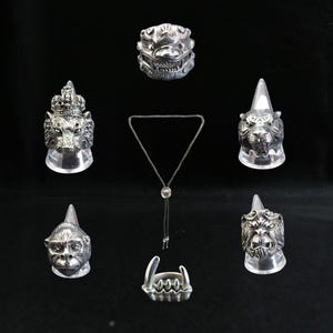 Peut inclure: Une collection de bagues et de pendentifs en métal argenté, chacun avec des motifs détaillés d'animaux ou de créatures mythiques. L'assortiment comprend une bague avec une tête de lion, un visage de singe et une bague avec un design denté. Un bolo tie est également inclus.