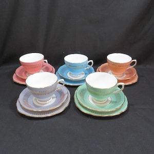 Könnte beinhalten: Fünf Vintage-Tassen und Untertassen in Pastellfarben: Rosa, Blau, Pfirsich, Lavendel und Mintgrün. Jede Tasse hat einen weißen Innenraum und goldfarbene Verzierungen. Die Untertassen haben ein passendes Farbschema mit einem dekorativen Muster.