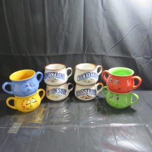 May include: Six ceramic soup mugs with cartoon faces. The mugs are stacked in pairs and feature the words "Minestrone", "Onion Soup", and "Chicken Soup".