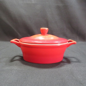 May include: A small, red ceramic casserole dish with a matching lid. The dish has two small handles and a rounded lid with a small knob. The interior is white. This kitchenware is suitable for baking and serving.