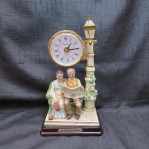 Pode incluir: Uma figurinha de porcelana de um casal mais velho sentado em um banco sob um poste de luz com um mostrador de relógio anexado. O mostrador do relógio tem algarismos romanos e uma moldura dourada. A base da figurinha é de madeira escura.