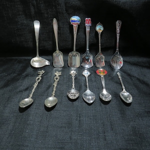 Könnte beinhalten: Eine Sammlung von 12 silbernen Löffeln mit verschiedenen Designs. Einige Löffel haben farbige Emaille-Designs, während andere verzierte Griffe haben. Die Löffel sind in einer Reihe auf einer schwarzen Oberfläche angeordnet.
