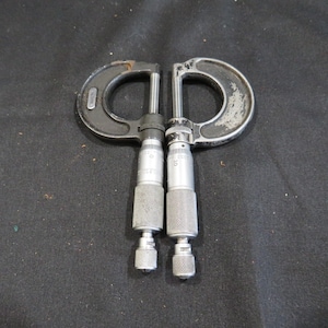 May include: Two vintage, used micrometer measuring tools. The tools are made of dark gray metal with a textured grip on the cylindrical portion. The C-shaped frame shows signs of wear. The tools are designed for precise measurements.