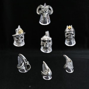 Può includere: Una collezione di anelli argentati esposti su supporti trasparenti. Gli anelli presentano vari design, tra cui teschi, una testa di ariete e una testa d'aquila. Gli anelli sono dettagliati e hanno un'estetica gotica o da motociclista.