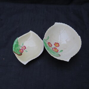 May include: Two leaf-shaped ceramic dishes. The dishes are off-white with a floral design featuring red and yellow flowers with green leaves. The dishes are on a dark background.