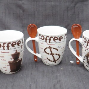 May include: Three white ceramic coffee mugs with brown coffee bean designs and matching brown spoons. The mugs feature different designs: one with a coffee pot, one with a dollar sign, and one with a bowl of coffee beans.