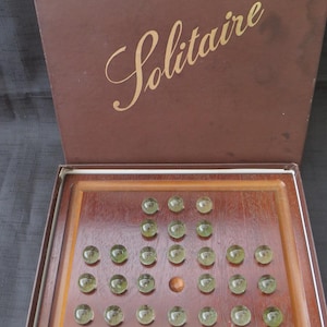 Peut inclure: Jeu de solitaire en bois avec une boîte et un plateau de jeu marron. Le couvercle de la boîte présente un motif de croix dorée et le mot "Solitaire" en écriture élégante. Le plateau contient des billes vertes, avec une bille marron centrale.