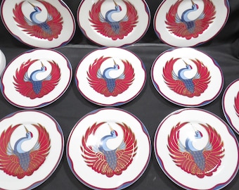 May include: A collection of white ceramic plates with a decorative crane design. The crane has red wings, blue body, and gold accents. The plates have a scalloped edge with a red and blue border. A set of decorative tableware.