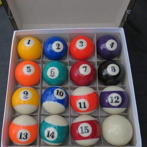 Könnte beinhalten: Ein vollständiger Satz Billardkugeln, nummeriert von 1 bis 15, plus eine weiße Kugel, angeordnet in einer weißen Pappschachtel. Die Kugeln sind in verschiedenen Farben gehalten, darunter Gelb, Blau, Rot, Lila, Orange, Grün und Schwarz. Die Zahlen sind weiß.