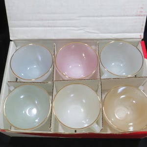 May include: A set of six pastel-coloured teacups with gold rims, arranged in a cardboard box. The cups are in shades of blue, pink, and green, with a white and tan cup. The cups have small handles.