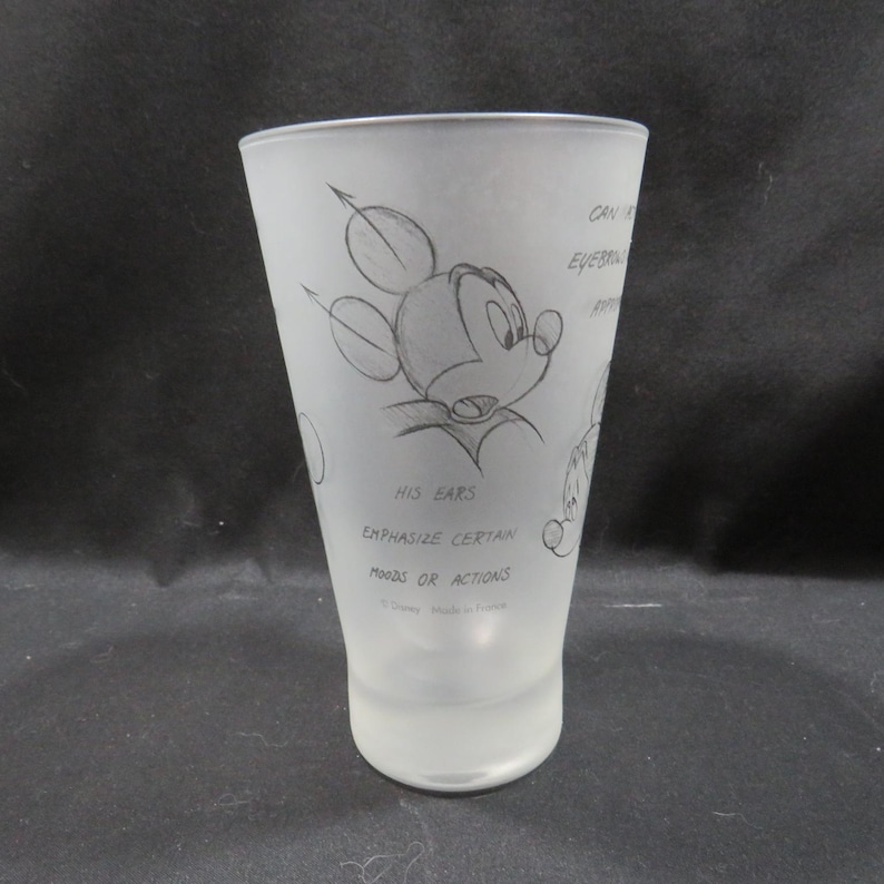 Op de afbeelding: Een mat glas met een schets van Mickey Mouse. De illustratie bevat Mickey's oren en de tekst "HIS EARS EMPHASIZE CERTAIN MOODS OR ACTIONS". Het glas is taps toelopend en heeft een gladde, matte afwerking.