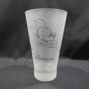 Op de afbeelding: Matte glazen tumbler met een schets van Mickey Mouse in beweging. Het ontwerp bevat het Disneyland Paris-logo in zwart. Het glas heeft een taps toelopende vorm en een gladde, matte afwerking, ideaal voor drankjes.