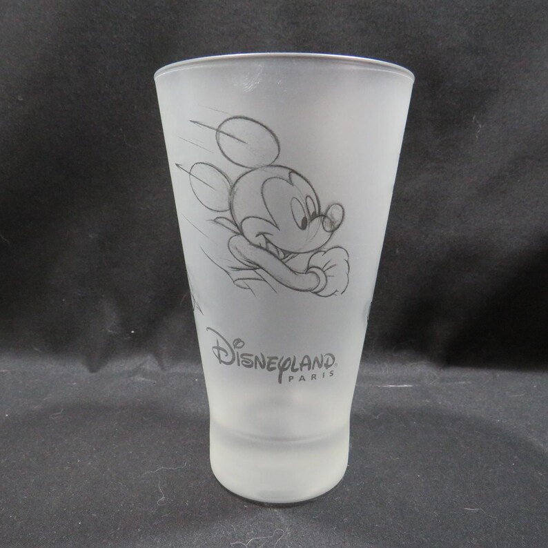 Op de afbeelding: Matte glazen tumbler met een schets van Mickey Mouse in beweging. Het ontwerp bevat het Disneyland Paris-logo in zwart. Het glas heeft een taps toelopende vorm en een gladde, matte afwerking, ideaal voor drankjes.