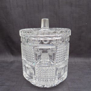 May include: A clear, cut-glass jar with a lid. The jar has a cylindrical shape with intricate geometric patterns and a small, rectangular handle on the lid. The glass reflects light, creating a sparkling effect. The jar is likely used for storage or display.