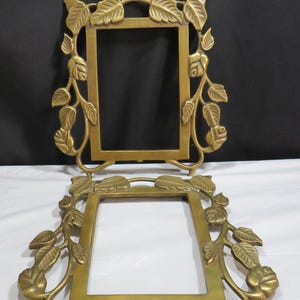 May include: Two brass picture frames with a rectangular aperture. The frames are decorated with a leaf and vine design. The frames are a warm gold colour and are displayed against a black and white background.