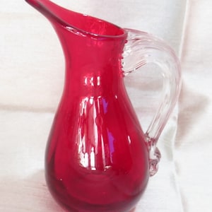 Puede incluir: Una pequeña jarra de vidrio roja con un asa de vidrio transparente. La jarra tiene un cuerpo redondeado y un labio ligeramente acampanado.