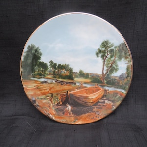 May include: Round decorative plate featuring a detailed landscape painting of a river scene with boats, trees, and a blue sky. The plate has a gold rim and a brown and orange color palette.