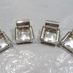 Könnte beinhalten: Vier kleine, silberfarbene Objekte in Stuhlform. Jeder Stuhl hat eine quadratische Sitzfläche und eine rechteckige Rückenlehne. Die Objekte sind reflektierend und scheinen aus Metall zu sein, möglicherweise Silber oder Chrom.