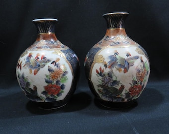 May include: Two antique Japanese ceramic vases. Each vase features a bulbous body, a narrow neck, and a flared rim. The vases are decorated with floral and bird motifs in shades of red, blue, and gold against a white and dark blue background.